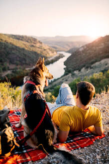 Back view of anonymous male hiker in yellowt shirt sitting on green hill with adorable dog in red collar and admiring picturesque scenery - ADSF49479