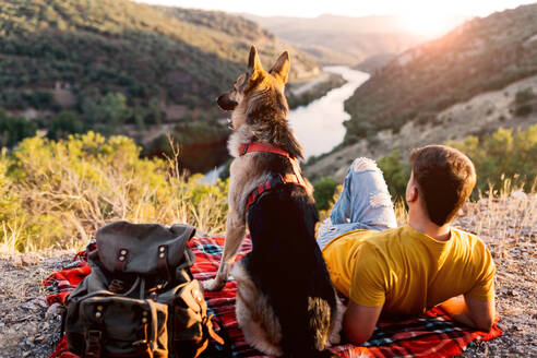 Back view of anonymous male hiker in yellowt shirt sitting on green hill with adorable dog in red collar and admiring picturesque scenery - ADSF49480