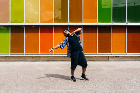 A latin man in sunglasses and headphones dancing against an urban building of multicolored panels. - ADSF49484