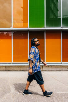 Latin young man with headphones walking in front of vivid panels, looking away. - ADSF49485