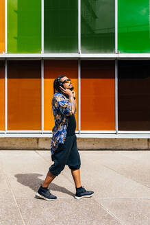 Latin man with braids and blue shirt and headphones walking by multicolored panels, looking away. - ADSF49486