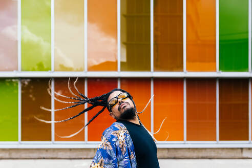 Latin man with long braids tossing his hair back, wearing sunglasses and a blue shirt against a colorful backdrop. - ADSF49488