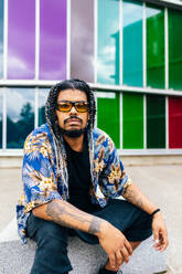 Latin man with braided hair and sunglasses seated against a multicolored windows, donning a blue floral shirt and tattoos on his arm. - ADSF49489