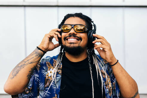 Latin man with braids and sunglasses enjoying music in black headphones against a white urban background. - ADSF49491