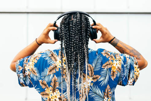 Latin man from behind of a latin man with detailed braids listening music with his black headphones, adorned with tattoos and wearing a floral blue shirt in front of a white wall background. - ADSF49492