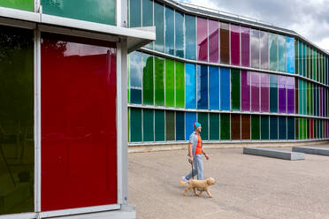 Man walking his dog past a multicolored glass building - ADSF49502