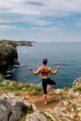 Mature man practicing yoga with a tranquil ocean backdrop, balancing on one foot while extending his arms in a scenic rocky location. - ADSF49511