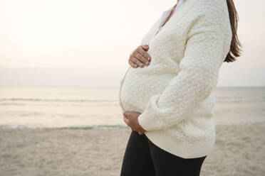 Schwangere Frau steht mit Händen auf dem Bauch am Strand - AAZF01238