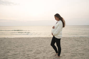 Glückliche schwangere Frau steht mit Händen auf dem Bauch am Strand - AAZF01239