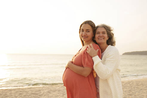 Glückliche schwangere Frau mit Mutter am Strand stehend - AAZF01252