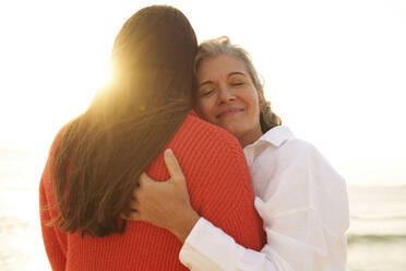 Smiling mother hugging daughter at beach - AAZF01264