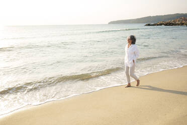 Glückliche reife Frau spaziert am Strand an einem sonnigen Tag - AAZF01276