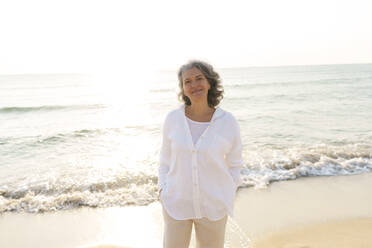 Lächelnde reife Frau mit Händen in den Taschen am Strand stehend - AAZF01279