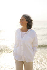Lächelnde reife Frau mit Händen in den Taschen am Strand stehend an einem sonnigen Tag - AAZF01280