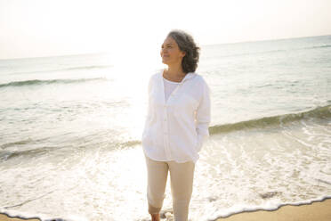 Smiling mature woman standing at beach on sunny day - AAZF01281