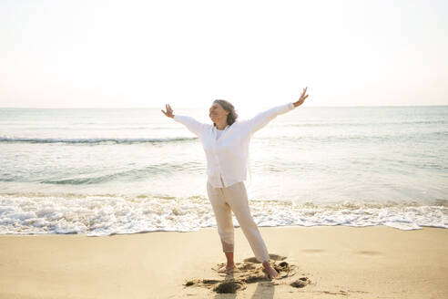 Carefree mature woman with arms outstretched standing at beach - AAZF01282