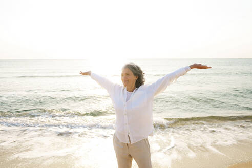 Smiling mature woman with arms outstretched standing at beach on sunny day - AAZF01283