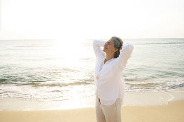 Smiling woman with hands behind head standing at beach on sunny day - AAZF01284
