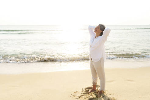 Smiling mature woman with hands behind head standing at beach - AAZF01285