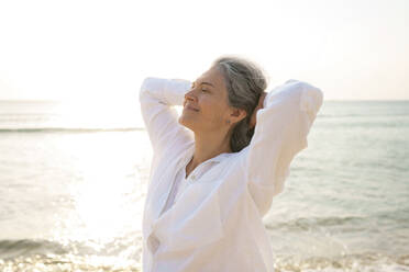 Smiling mature woman standing with hands behind head in front of sea - AAZF01286