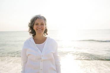 Smiling mature woman standing in front of sea at beach - AAZF01288