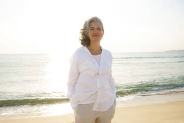 Smiling mature woman standing in front of sea at beach on sunny day - AAZF01289