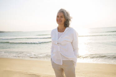 Smiling mature woman standing at beach - AAZF01290