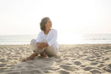 Smiling mature woman sitting on sand at beach - AAZF01291