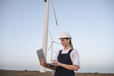 Lächelnder Ingenieur mit Laptop in der Nähe von Windkraftanlagen - ALKF00835