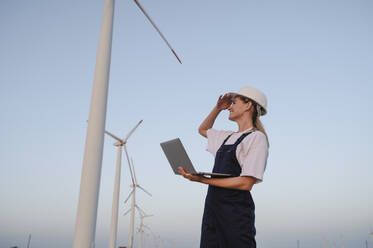 Glücklicher Ingenieur mit Augenschutz und Laptop in der Nähe von Windkraftanlagen - ALKF00836