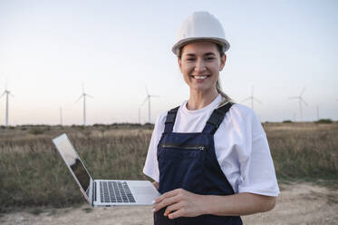 Glücklicher Ingenieur mit Schutzhelm und Laptop vor einer Windkraftanlage bei Sonnenuntergang - ALKF00839