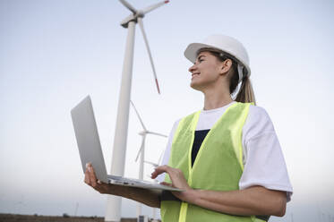 Glücklicher Ingenieur mit Laptop vor einer Windkraftanlage - ALKF00854