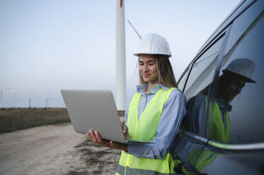 Ein lächelnder Ingenieur arbeitet an einem Laptop in der Nähe eines Autos auf einem Windkraftanlagenfeld - ALKF00859