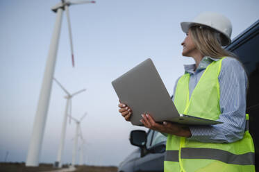 Ein lächelnder Ingenieur steht mit einem Laptop in der Nähe eines Autos auf einem Windkraftanlagenfeld - ALKF00860