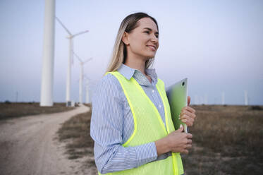 Smiling engineer holding laptop and standing in front of wind turbines - ALKF00866