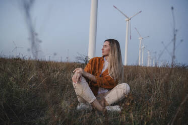 Nachdenkliche Frau sitzt vor Windrädern auf einem Feld - ALKF00874