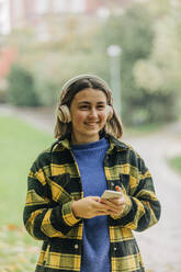 Lächelndes Teenager-Mädchen, das ein Smartphone in der Hand hält und über drahtlose Kopfhörer im Park Musik hört - VSNF01463