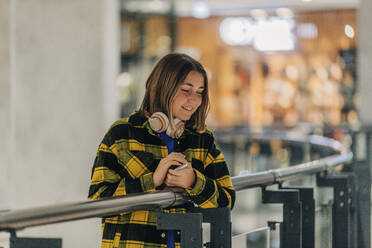 Lächelndes Teenager-Mädchen mit drahtlosen Kopfhörern, das in der Nähe eines Geländers in einem Einkaufszentrum steht - VSNF01474