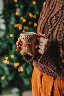 Woman holding cup of coffee with illuminated lights in background at home - VSNF01481