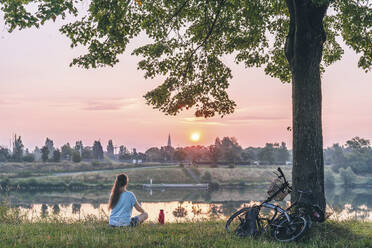 Mature woman sitting on grass near river and watching sunrise - TILF00002