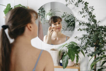 Woman applying cream on face in front of mirror - TILF00003