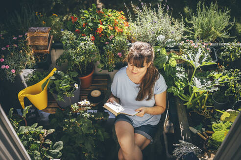 Woman sitting and reading book near plants in balcony - TILF00006