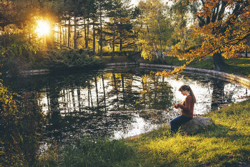 Frau sitzt auf einem Felsen in der Nähe eines Teiches im Herbstpark - TILF00018