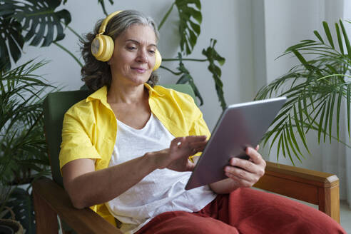 Frau mit drahtlosen Kopfhörern und Tablet-PC auf einem Sessel zu Hause - AAZF01304