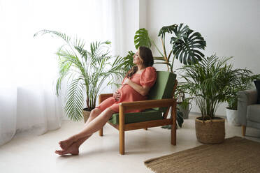 Pregnant woman relaxing on armchair near plants at home - AAZF01335