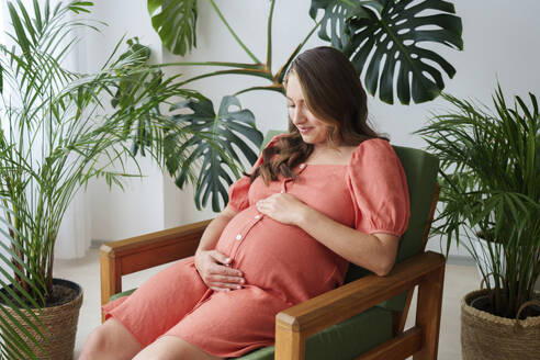 Smiling pregnant woman looking at belly sitting on armchair at home - AAZF01337