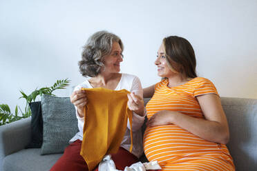 Schwangere Frau sieht Mutter mit Babykleidung auf dem Sofa sitzend zu Hause an - AAZF01344