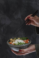 Hände einer Frau, die eine Schüssel mit veganer Tom kha kai-Suppe mit Tofu, Tomaten, Salat, Reisnudeln, Sesam und Frühlingszwiebeln isst - EVGF04421