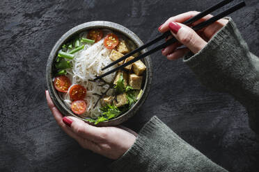 Hands of woman eating bowl of vegan Tom kha kai soup with tofu, tomatoes, salad, rice noodles, sesame seeds and scallion - EVGF04424