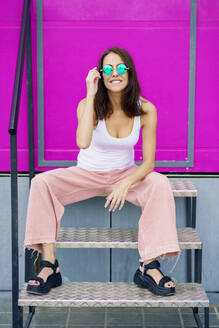 Lächelnde Frau mit Sonnenbrille, die auf einer Treppe vor einem rosa Hintergrund sitzt - JSMF02944